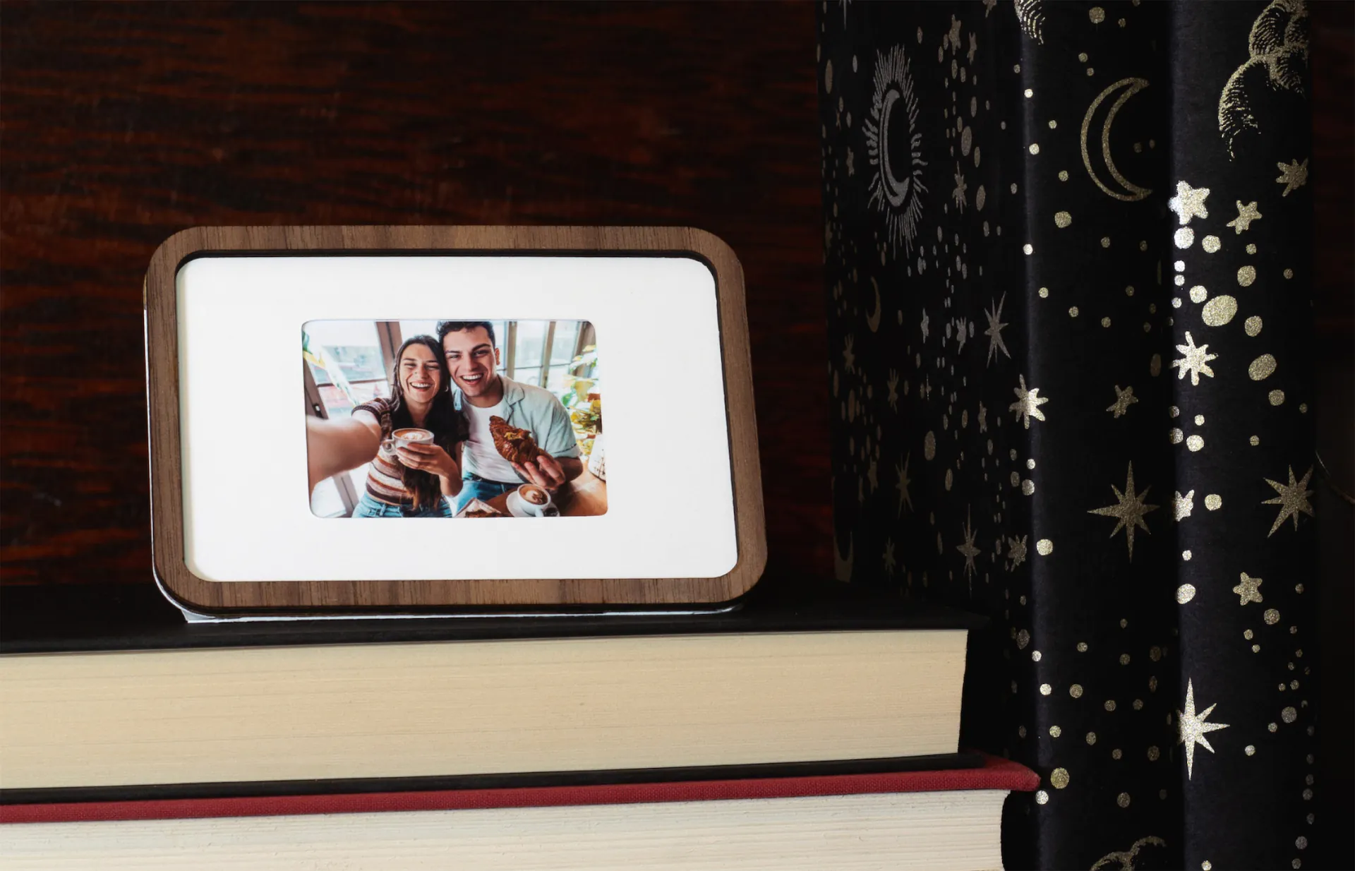 A walnut Pop-up Frame with a photo of two friends at a café, displayed on a stack of books